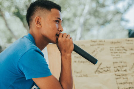 A teen boy is speaking on a mic and delivering some speech, a whiteboard can be seen nearby with something written on it.