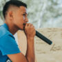 A teen boy is speaking on a mic and delivering some speech, a whiteboard can be seen nearby with something written on it.