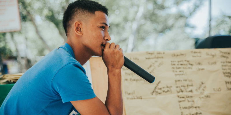 A teen boy is speaking on a mic and delivering some speech, a whiteboard can be seen nearby with something written on it.