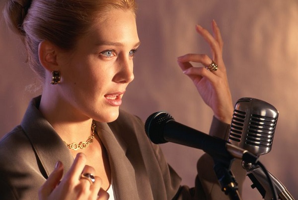 A woman is giving speech on a mic in front of the people.